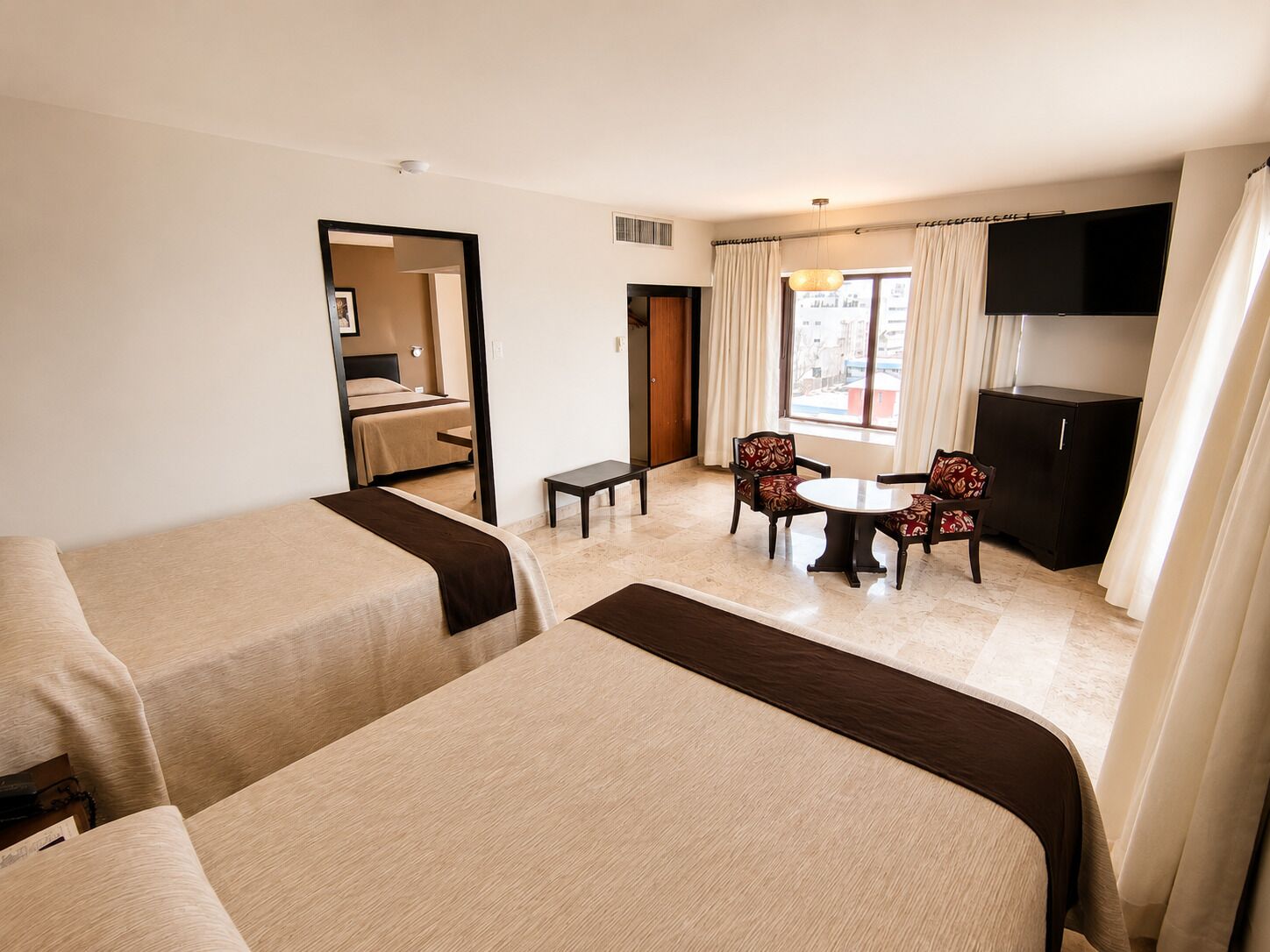Une chambre d’hôtel avec deux lits, une petite table, deux chaises, un téléviseur et une fenêtre avec des rideaux.