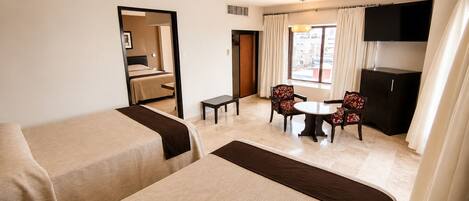 Une chambre d’hôtel avec deux lits, une petite table, deux chaises, un téléviseur et une fenêtre avec des rideaux.