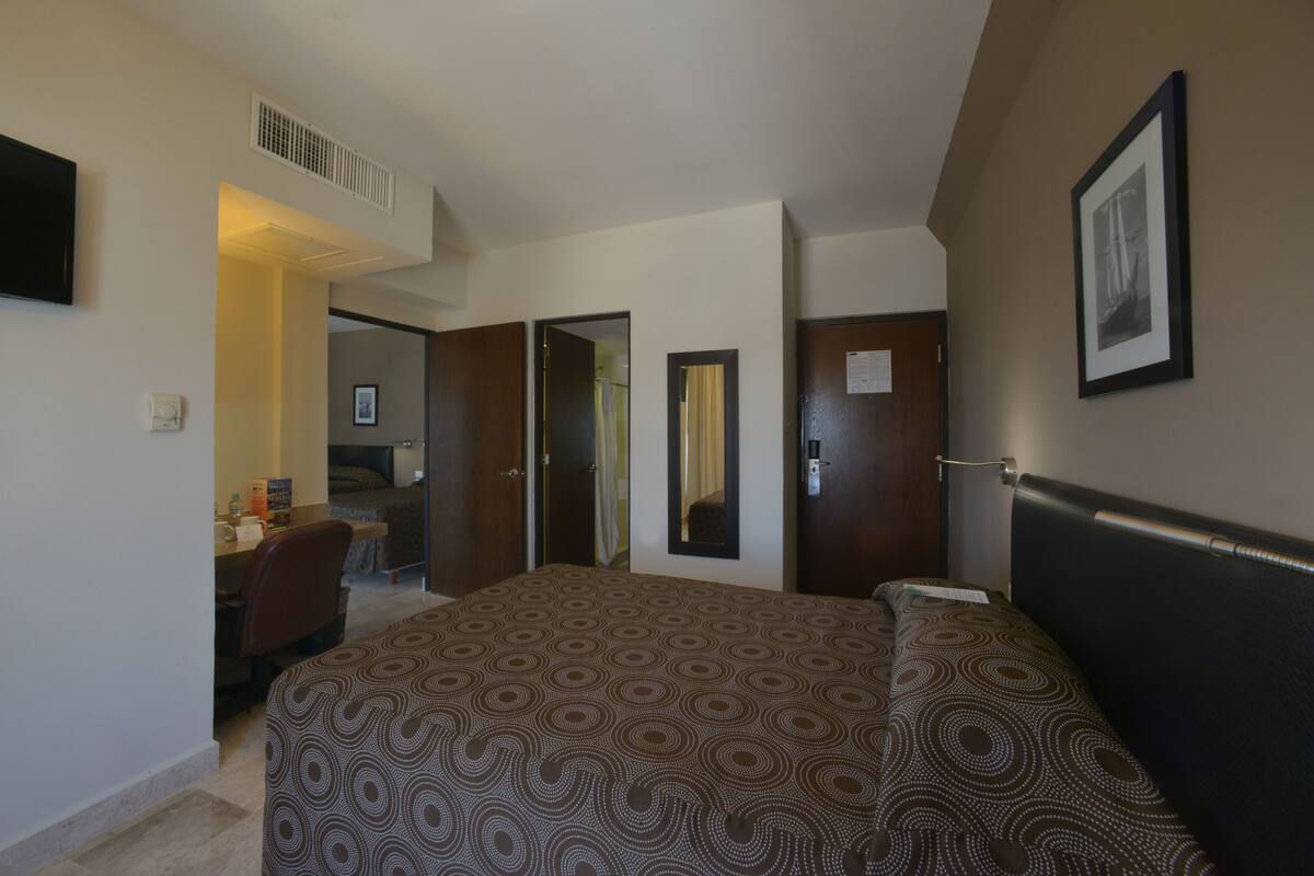 A hotel room with a bed, a desk, a chair, a mirror, and a framed picture on the wall.
