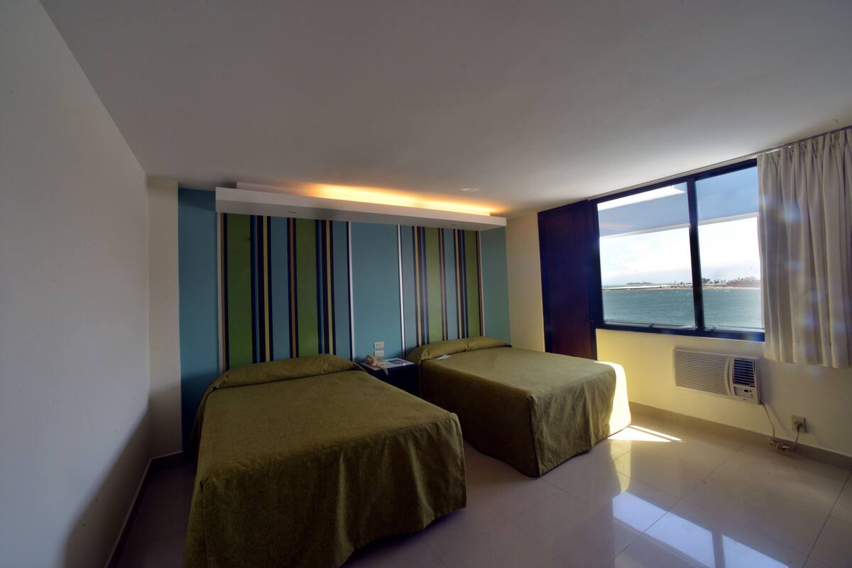 A hotel room with two single beds, a nightstand, a window with a view of the sea, and a striped wall design.