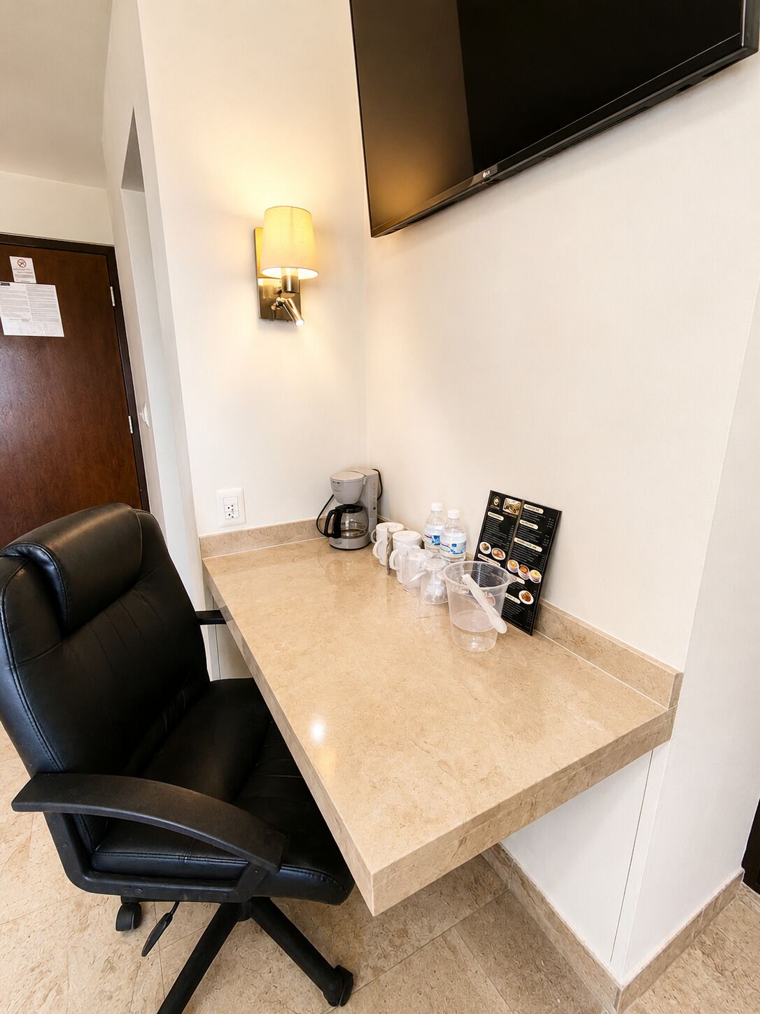 A hotel room with a flat-screen TV mounted on the wall, a desk with a chair, a coffee maker, and a menu board.