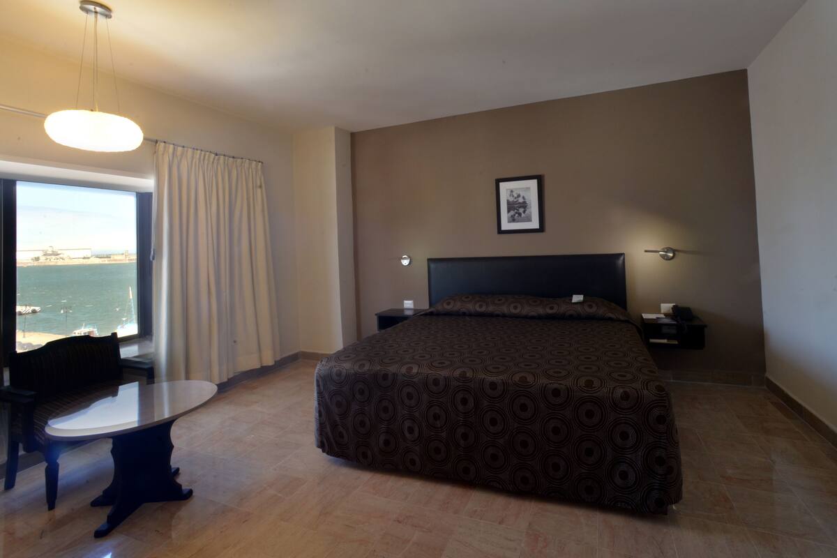 A hotel room with a bed, a desk, a chair, a window with curtains, a framed picture on the wall, and a view of the sea.