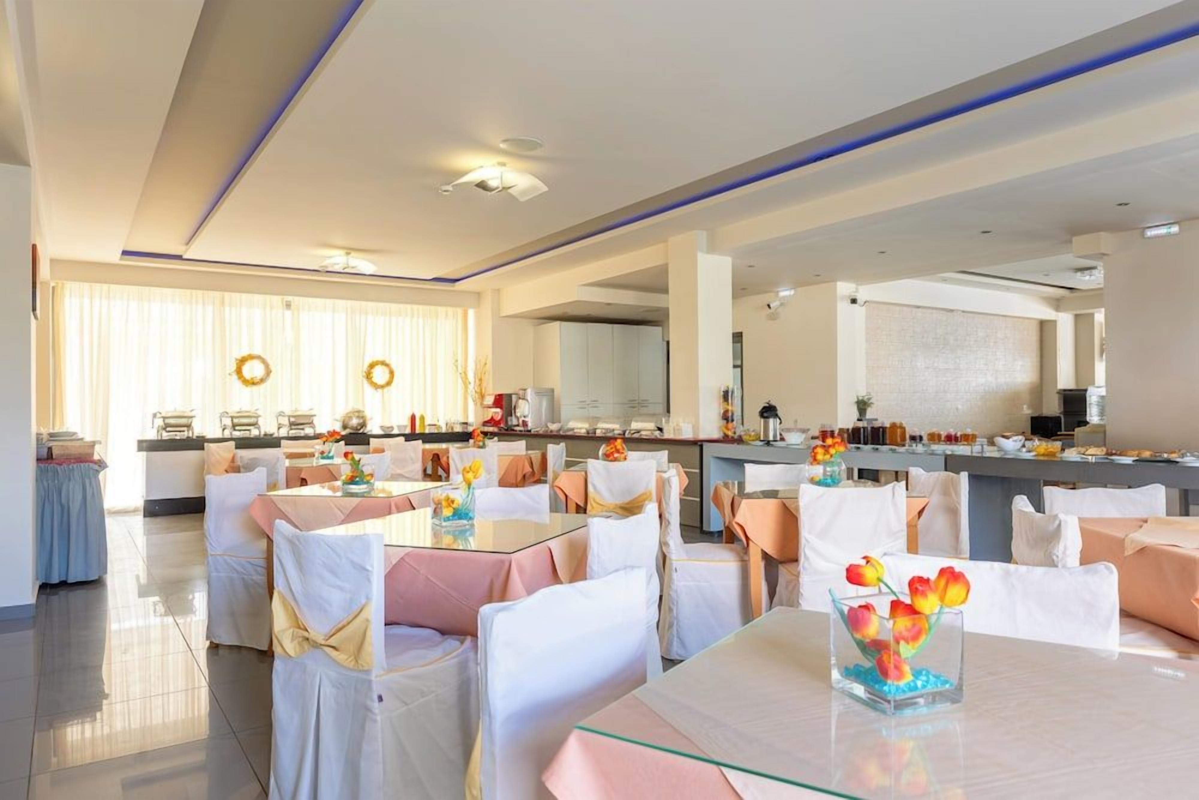 Ein Hotelrestaurant mit mehreren Tischen und Stühlen, einer Küche im Hintergrund und einem hellen, freundlichen Ambiente.