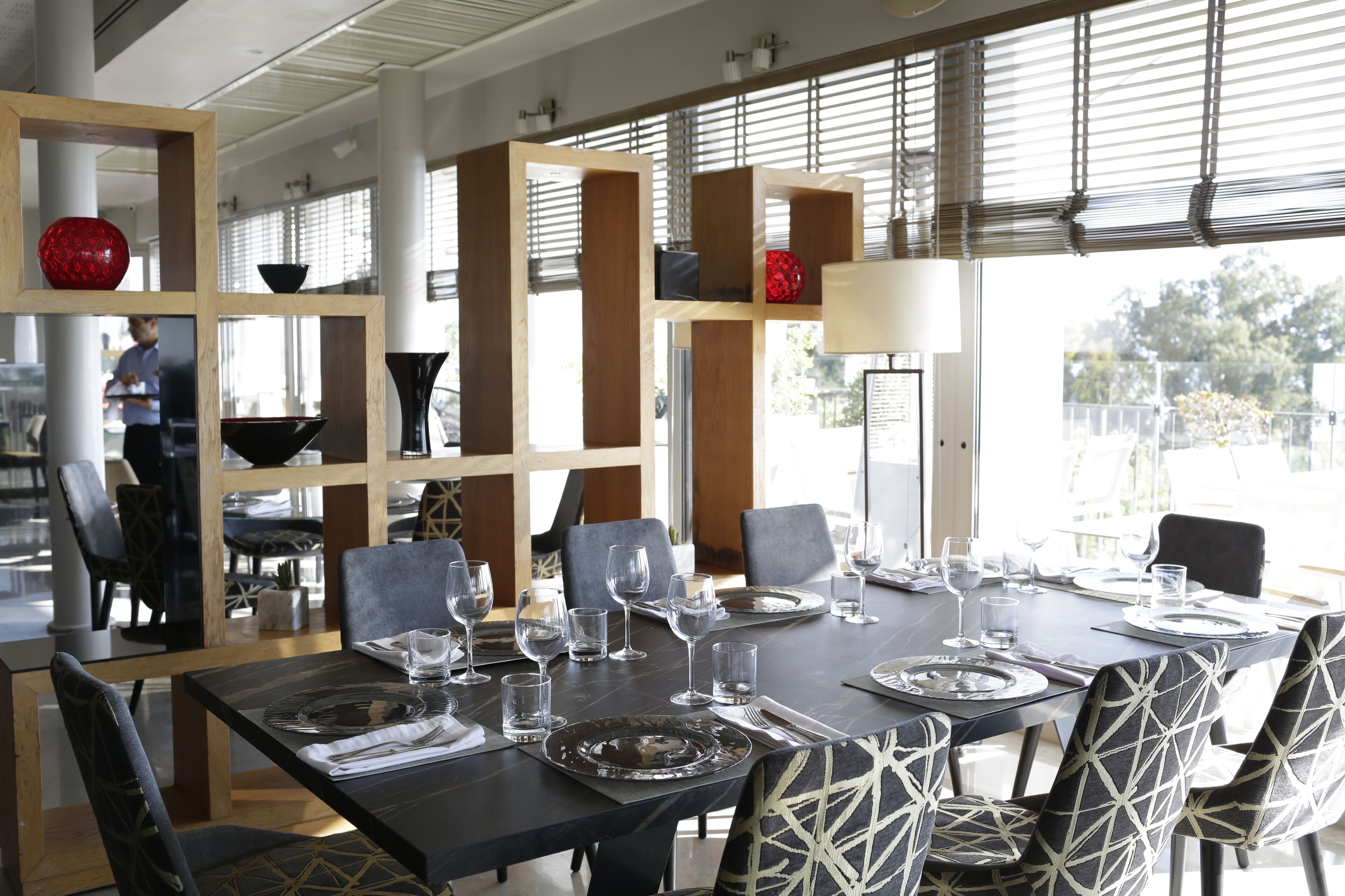 A dining area with a large table set for six, surrounded by chairs with geometric patterns. There are multiple wine glasses and plates on the table, and a red spherical object on a shelf.