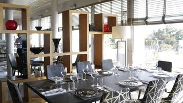 Uma sala de jantar com uma mesa grande posta para seis pessoas, rodeada por cadeiras com padrões geométricos. Há vários copos de vinho e pratos na mesa, e um objeto esférico vermelho em uma prateleira.