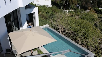 Une piscine sur le toit, agrémentée d’un parasol beige, entourée d’une végétation luxuriante et avec un bâtiment blanc en arrière-plan.