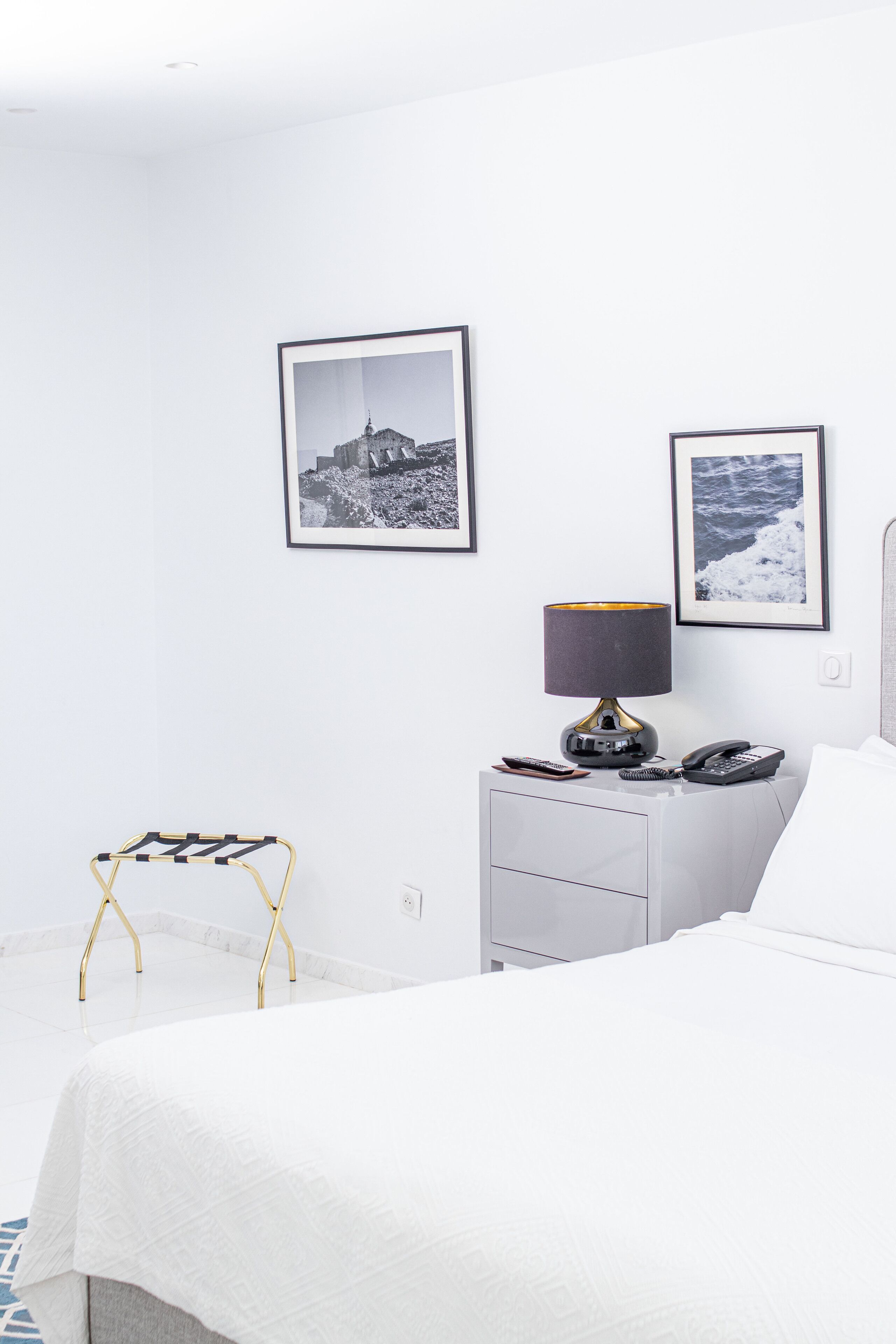 A hotel room with a bed, a bedside table with a lamp, a phone, and a picture frame on the wall.