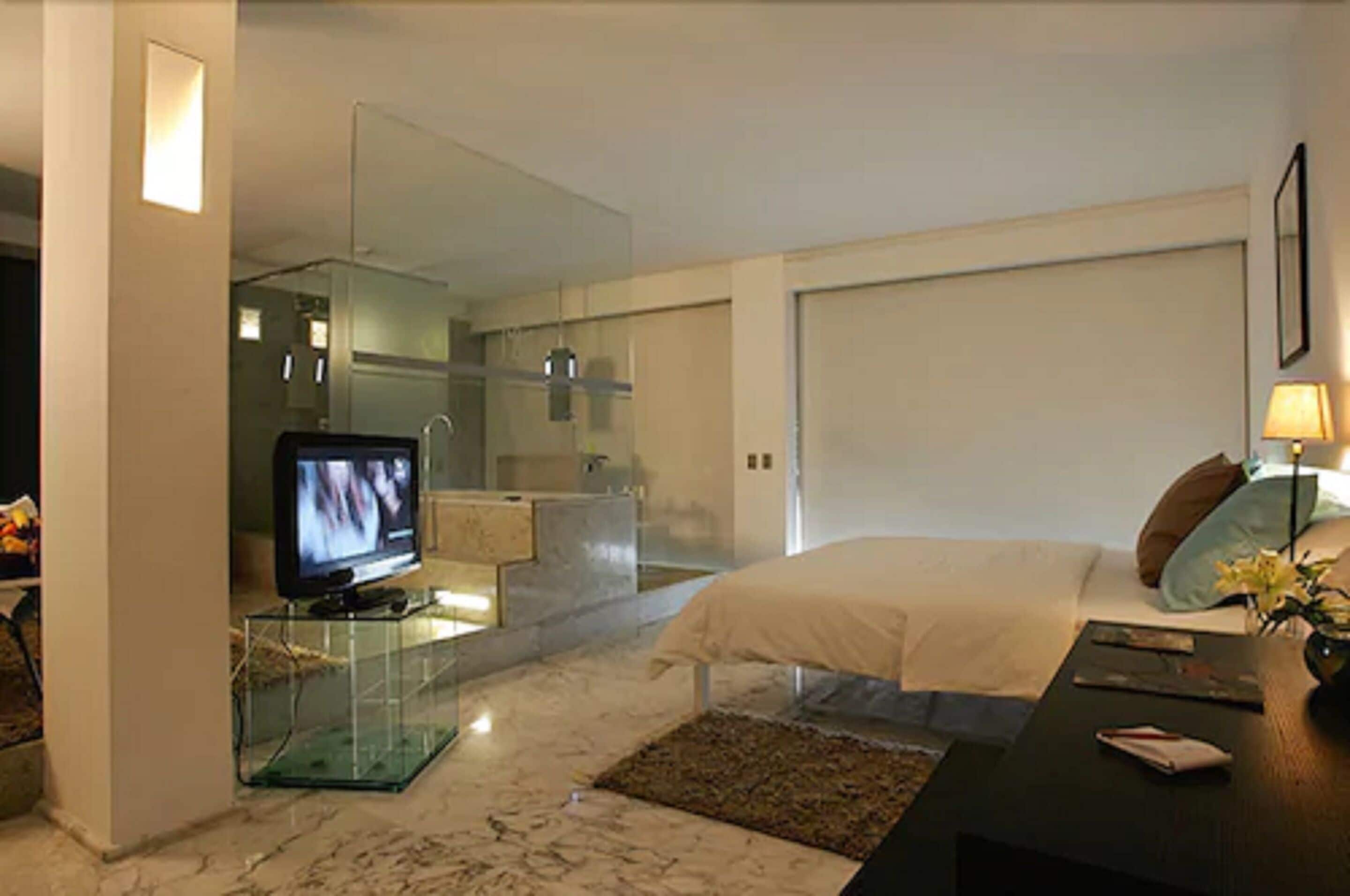 A modern bedroom with a bed, a television on a glass table, a bathroom with a glass shower enclosure, and a nightstand with a lamp.