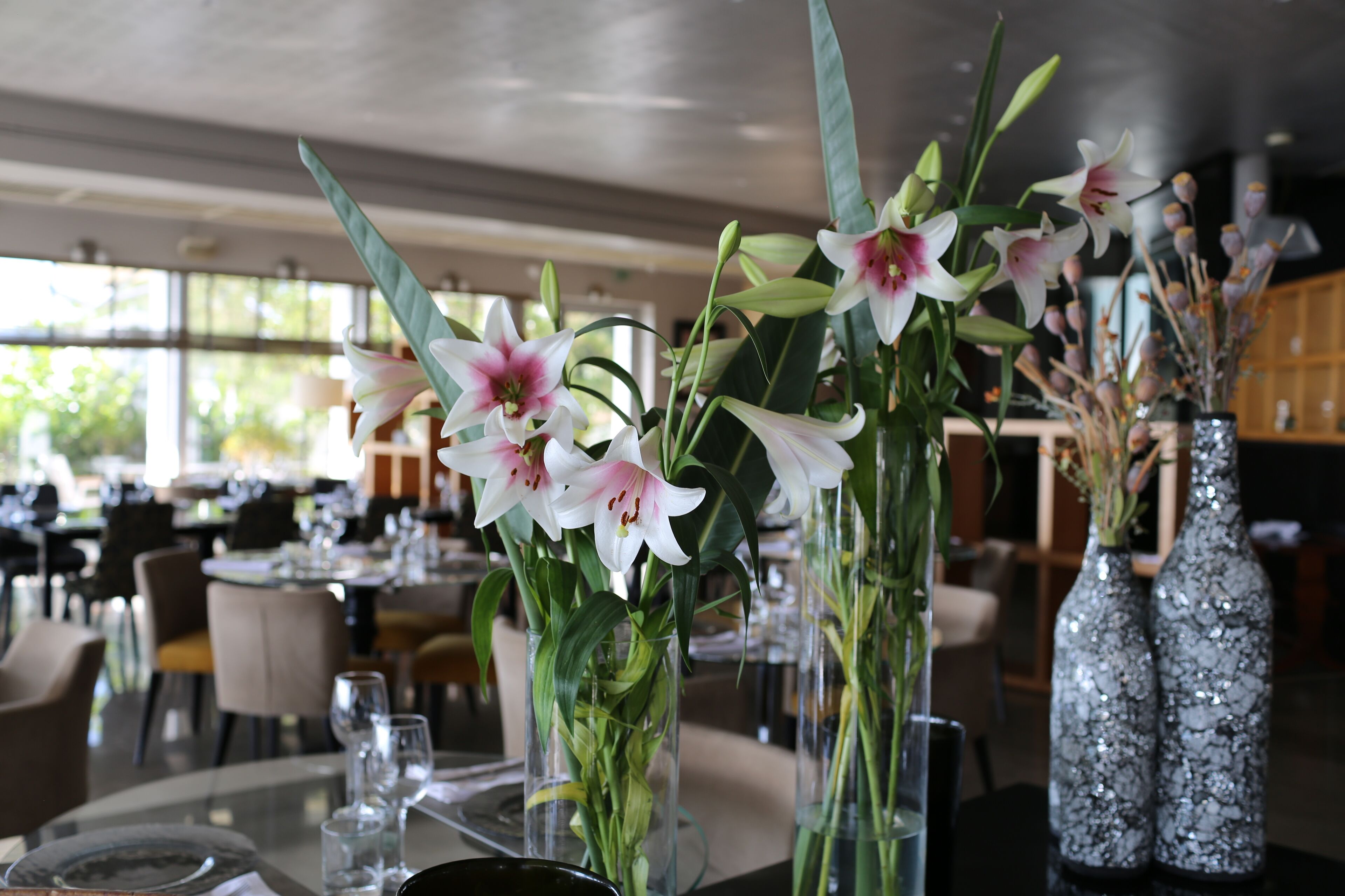Interior de un restaurante con mesas preparadas para comer, destacando jarrones con flores y cristalería transparente.