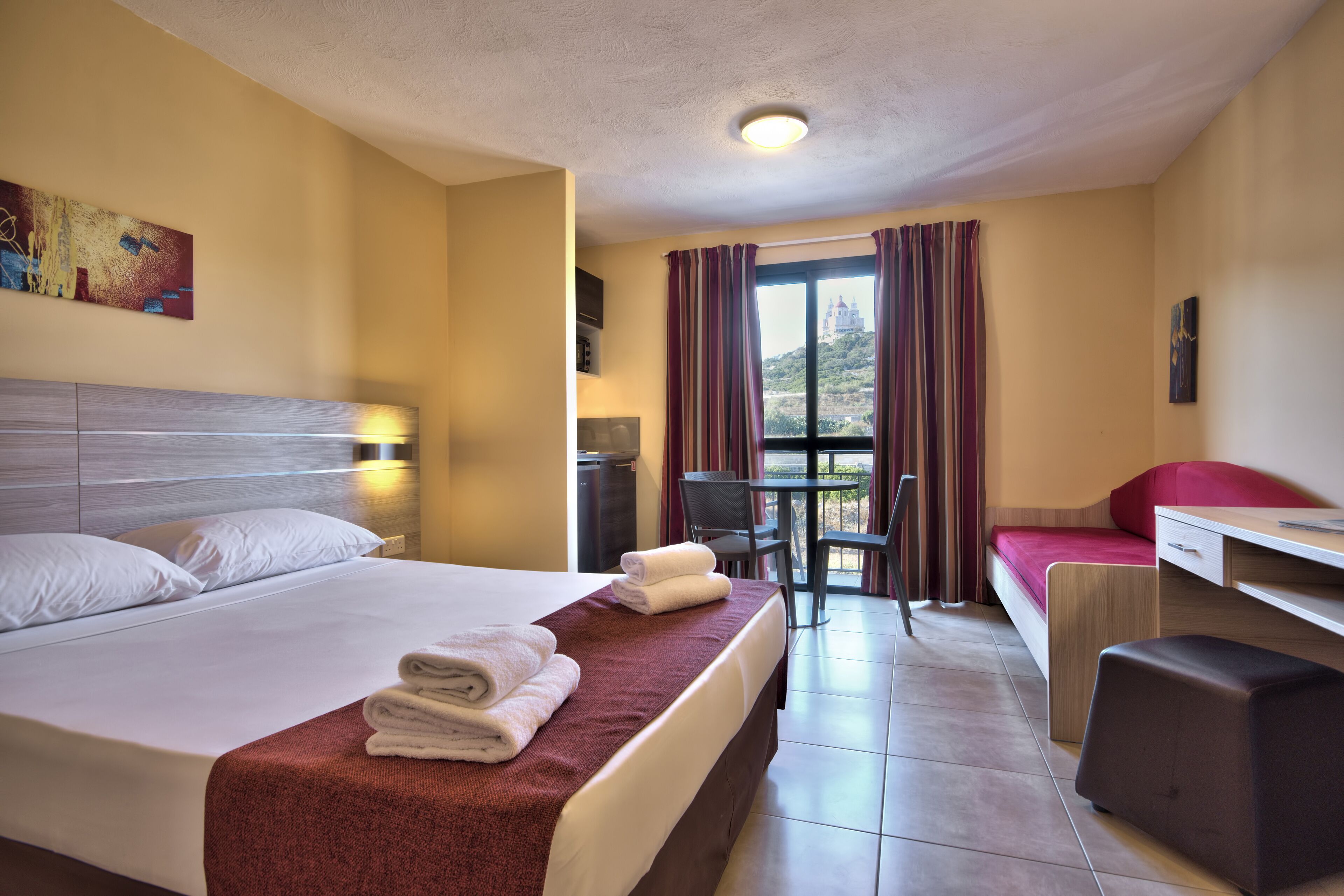 Habitación de hotel con cama, escritorio, silla, mesita y vista a un edificio a través del ventanal.