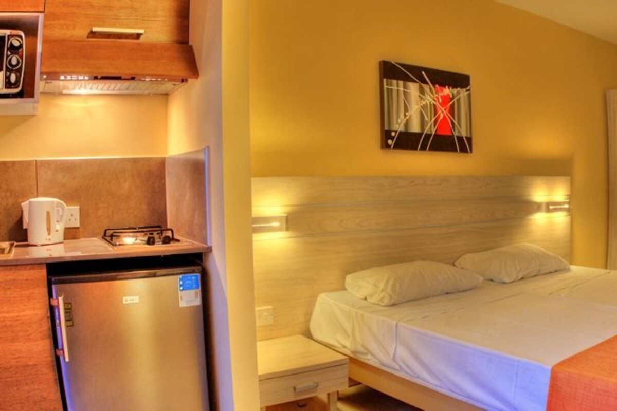 A compact hotel room with a kitchenette, a bed with white linens, a nightstand, and a wall-mounted artwork.