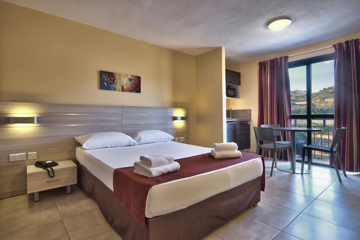 A hotel room with a large bed, bedside lamps, a desk with a chair, a view of the outdoors, and a painting on the wall.