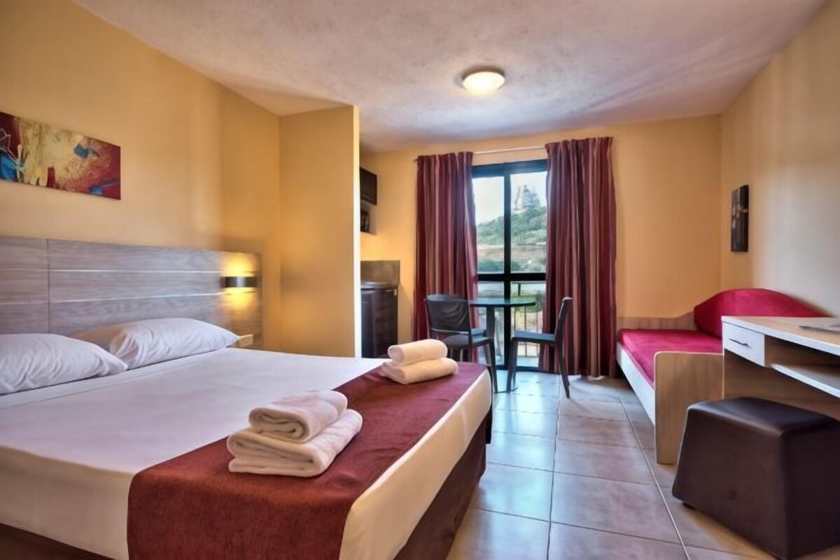A hotel room with a bed, a desk, a chair, a small table, and a window with curtains.