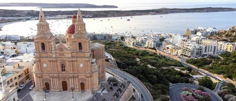 A historic church with a prominent bell tower and a clock, surrounded by a coastal town with modern buildings and a clear view of the sea.