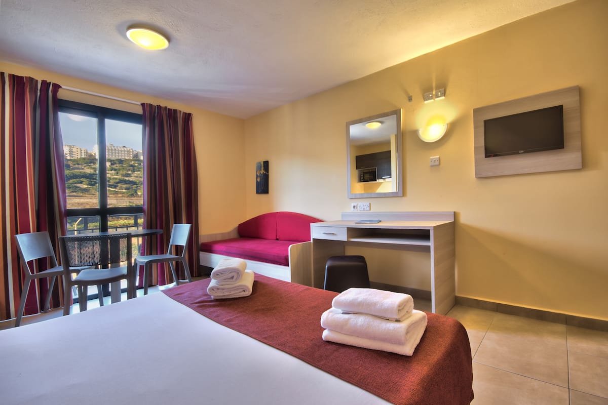 A hotel room with a bed, a desk, a chair, a TV, and a view of the outside.