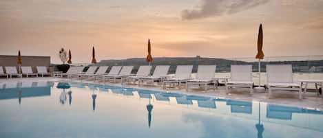 Zona junto a la piscina con sillones de descanso y sombrillas, una piscina cristalina y una vista del paisaje de fondo.
