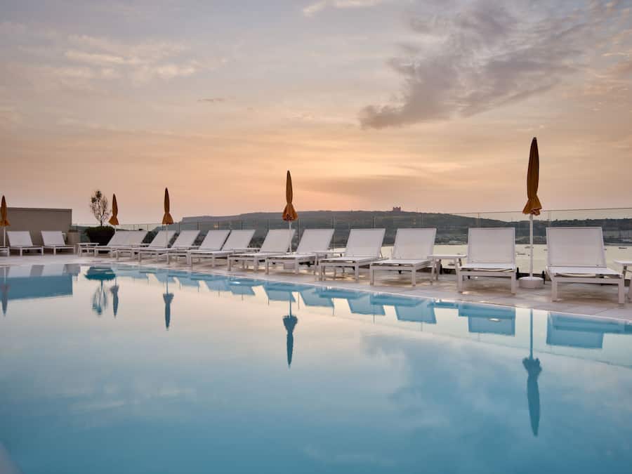 Zona junto a la piscina con sillones de descanso y sombrillas, una piscina cristalina y una vista del paisaje de fondo.