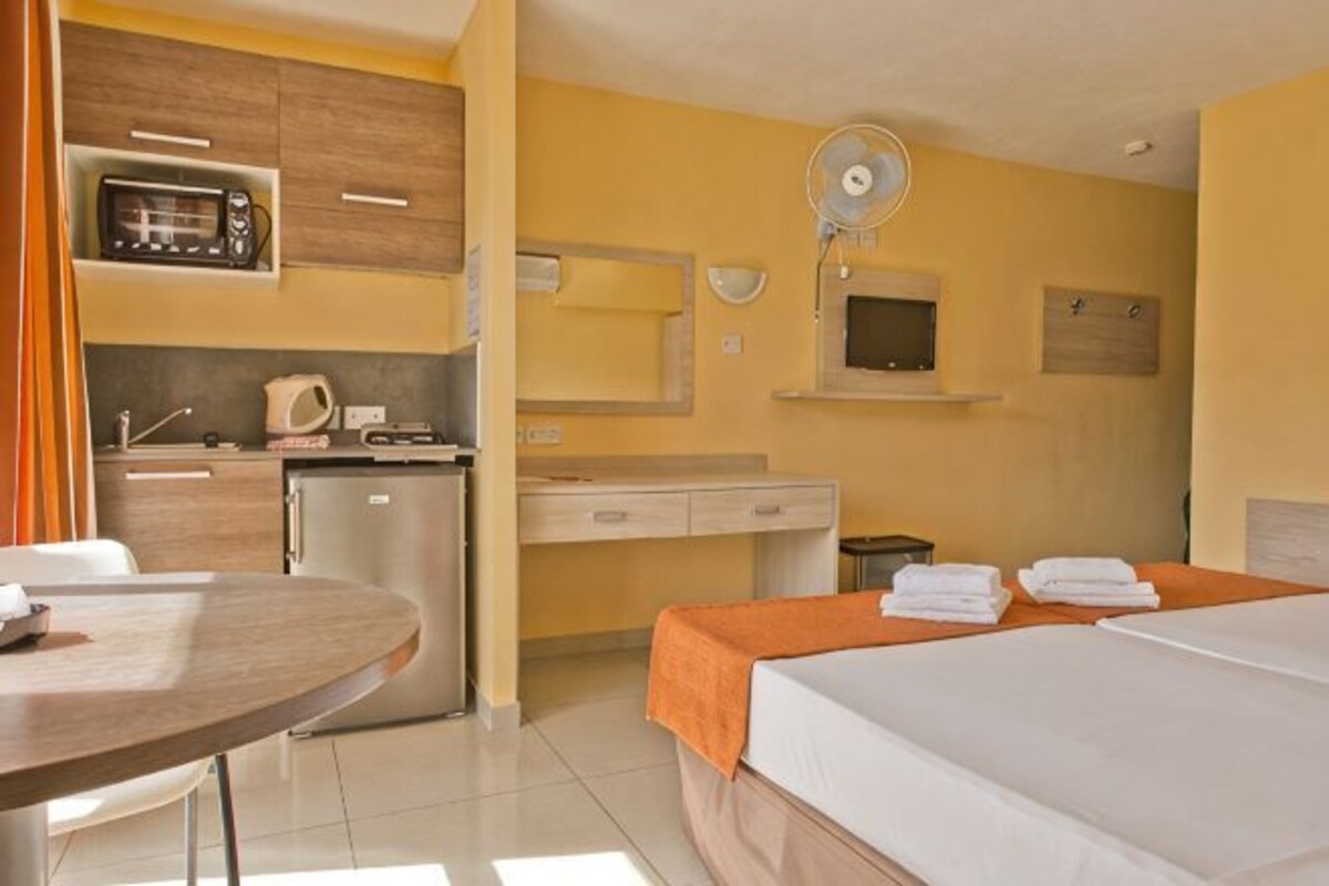A hotel room with a bed, a small kitchenette, a dining table, and a TV.