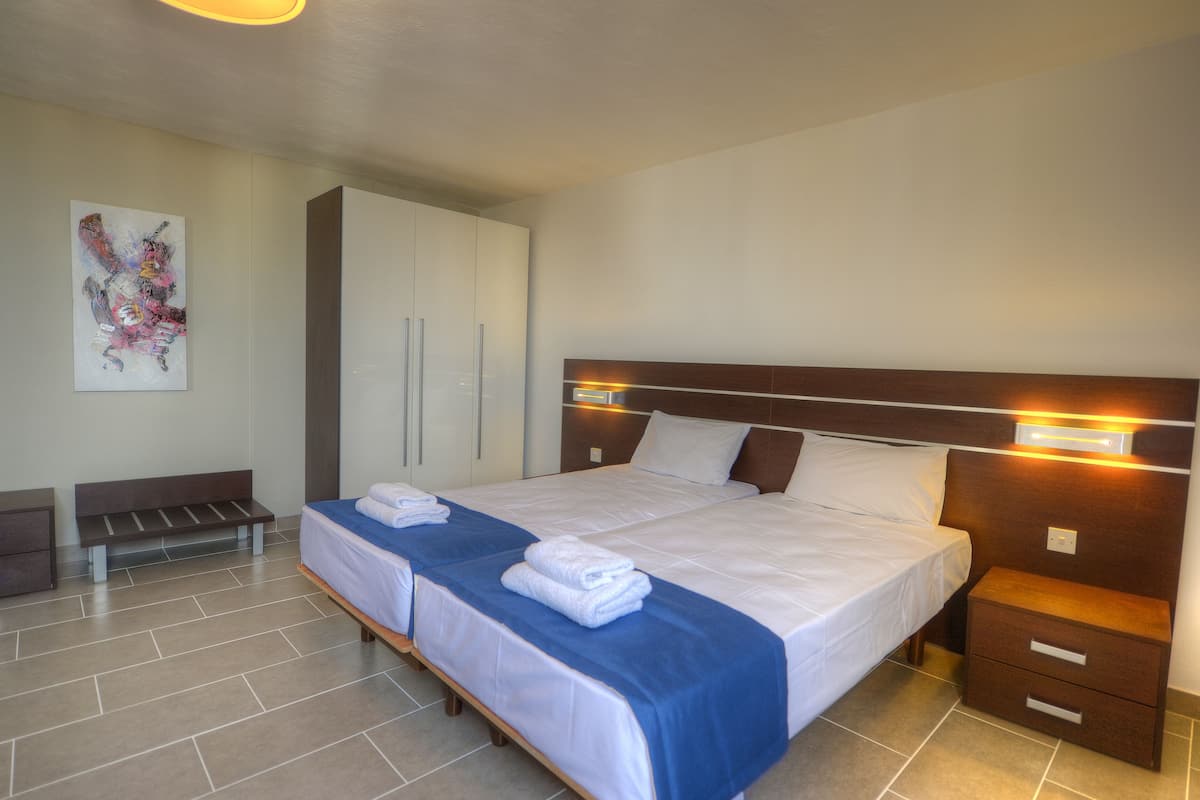 A double bed with a wooden headboard, two bedside tables, a bench, and a wall-mounted artwork.