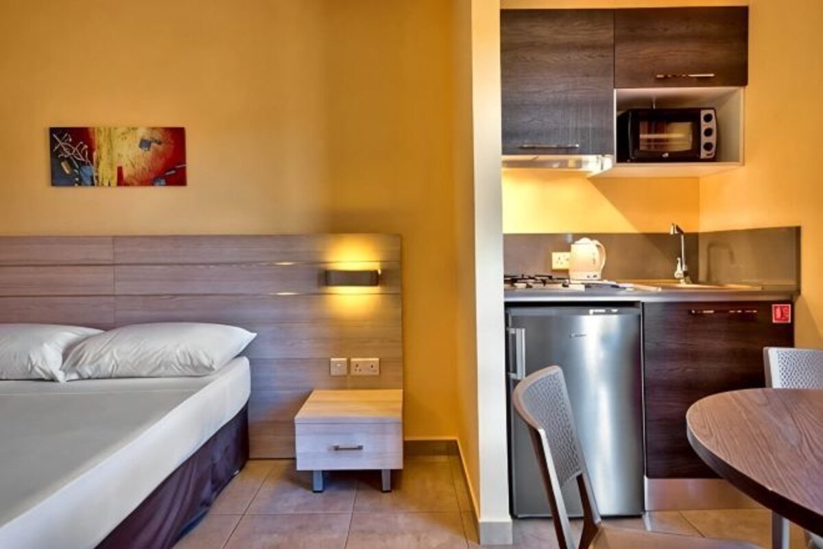 A compact hotel room with a bed, bedside table, and a kitchenette area including a microwave, stove, and sink.