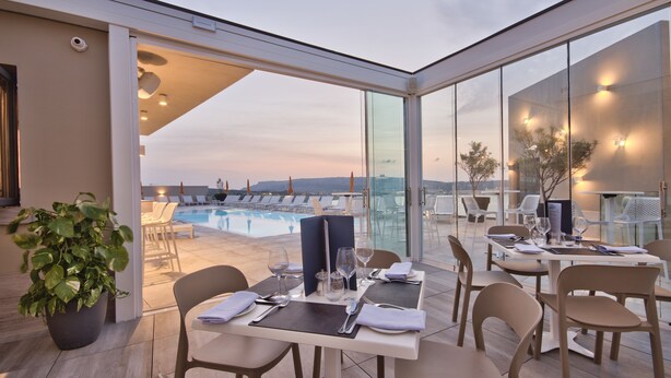 A restaurant with a glass-enclosed patio overlooking a pool and lounge area. - Luna Holiday Complex (Mellieha)