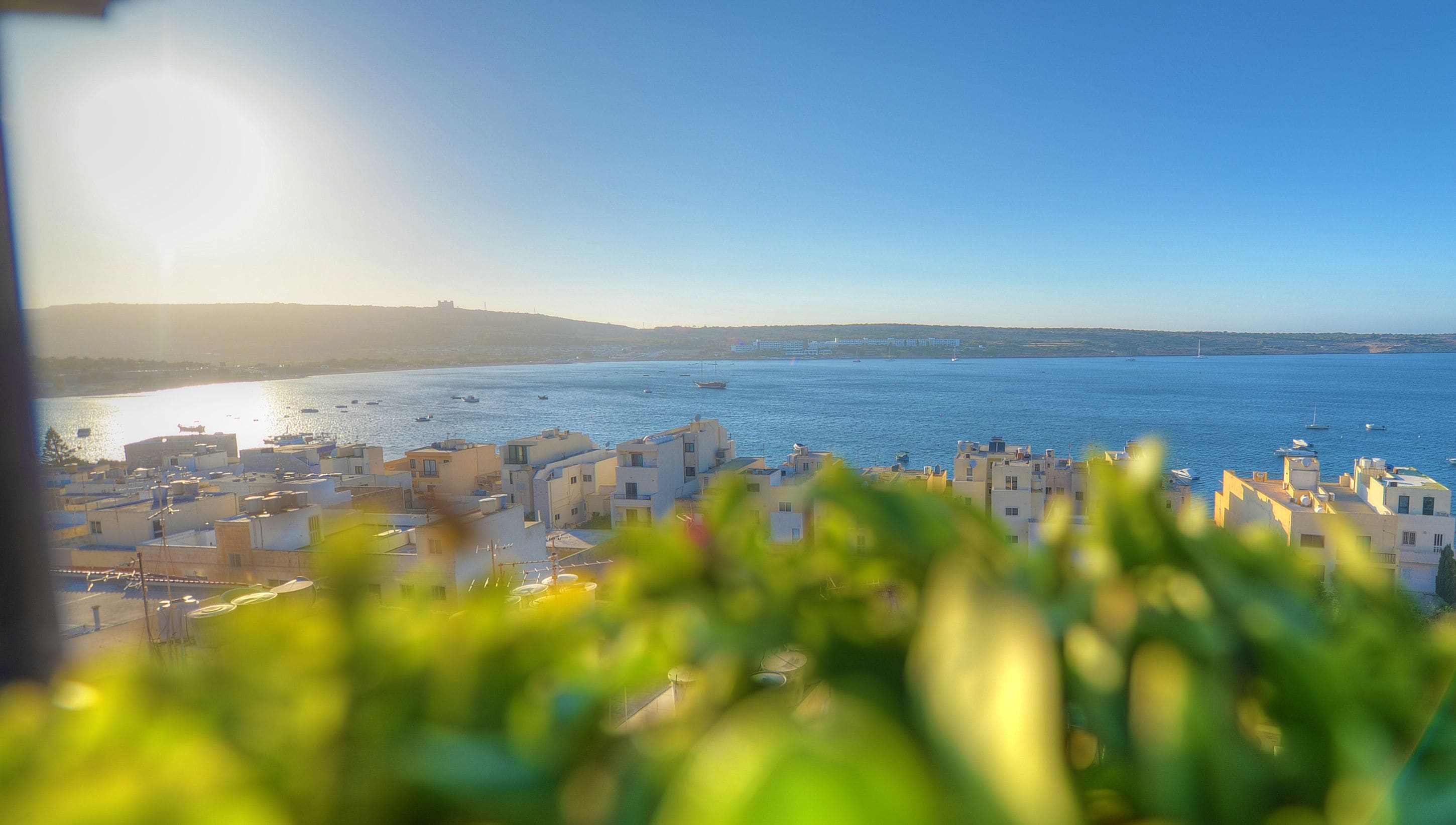 A coastal cityscape with buildings overlooking the sea, boats, and a clear sky.