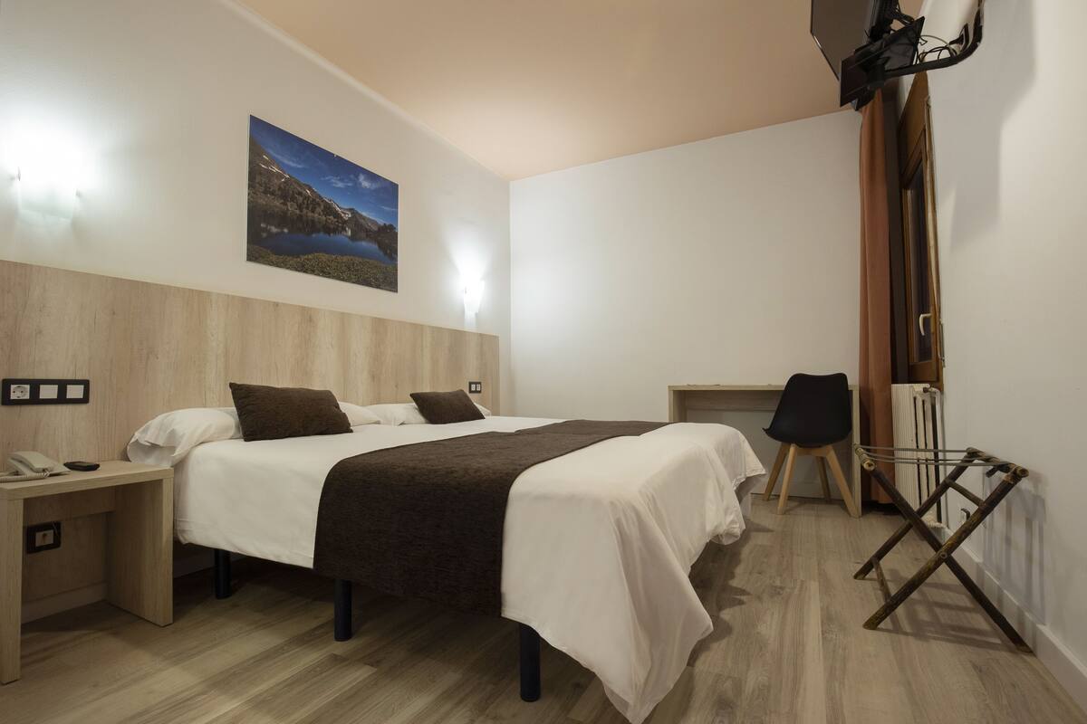 Moderni hotellihuone, jossa on puinen sängynpääty, suuri sänky valkoisilla ja ruskeilla vuodevaatteilla, työpöytä puhelimineen sekä seinälle asennettu televisio.