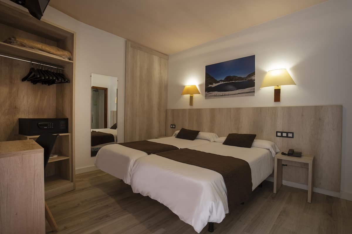 Hotellihuone, jossa on kaksi sänkyä, puulattia, vaatekaappi, yöpöytä lampun kanssa ja taulu seinällä.
