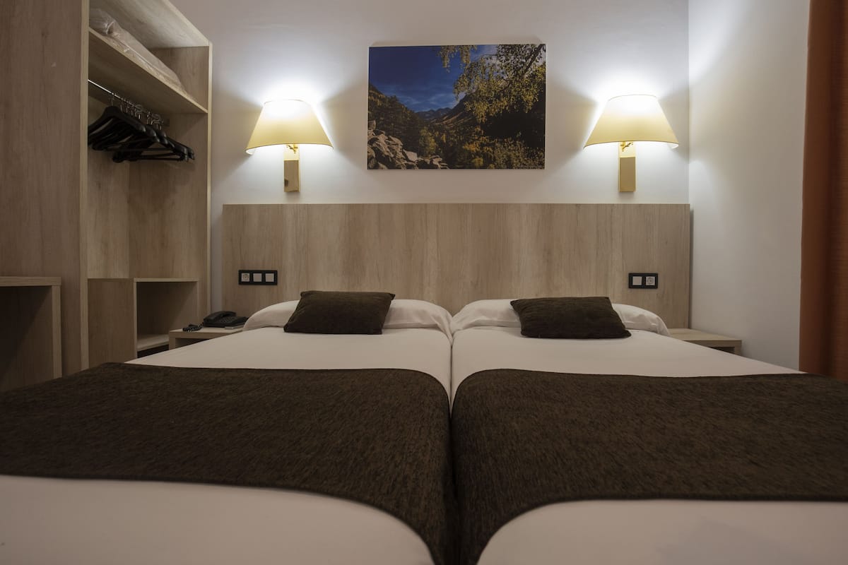 Hotellihuone, jossa on kaksi sänkyä, puinen vaatekaappi, yöpöytä lampun kanssa ja taulu seinällä.