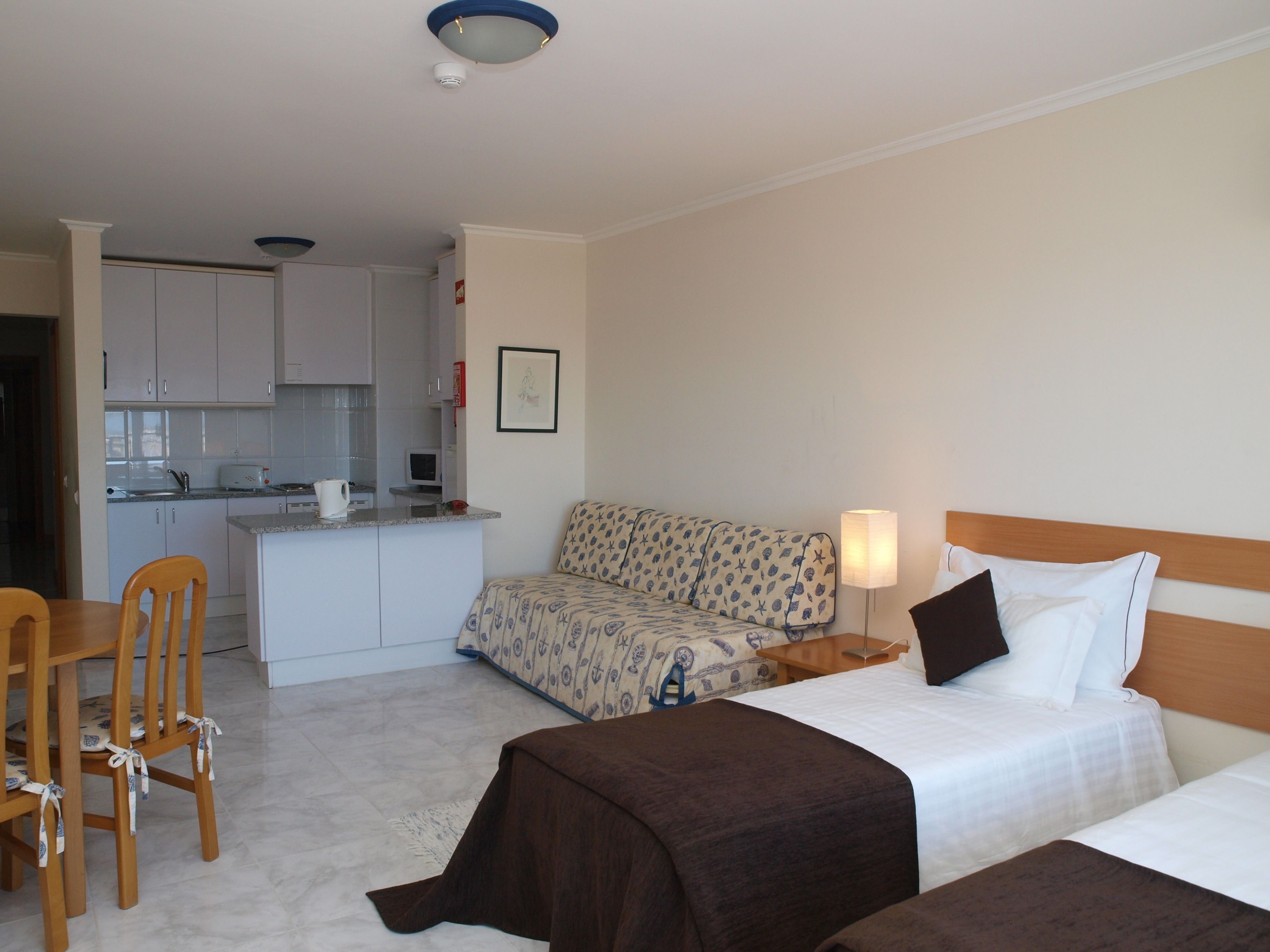 A hotel room with a bed, bedside table, lamp, and a kitchenette area.
