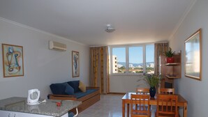 A modern apartment with a kitchen, dining area, and living space. It features a sofa, a dining table with chairs, a kitchen island with bar stools, and a large window offering a view of buildings and mountains. - Apartamentos Jardins da Rocha (Portimão)