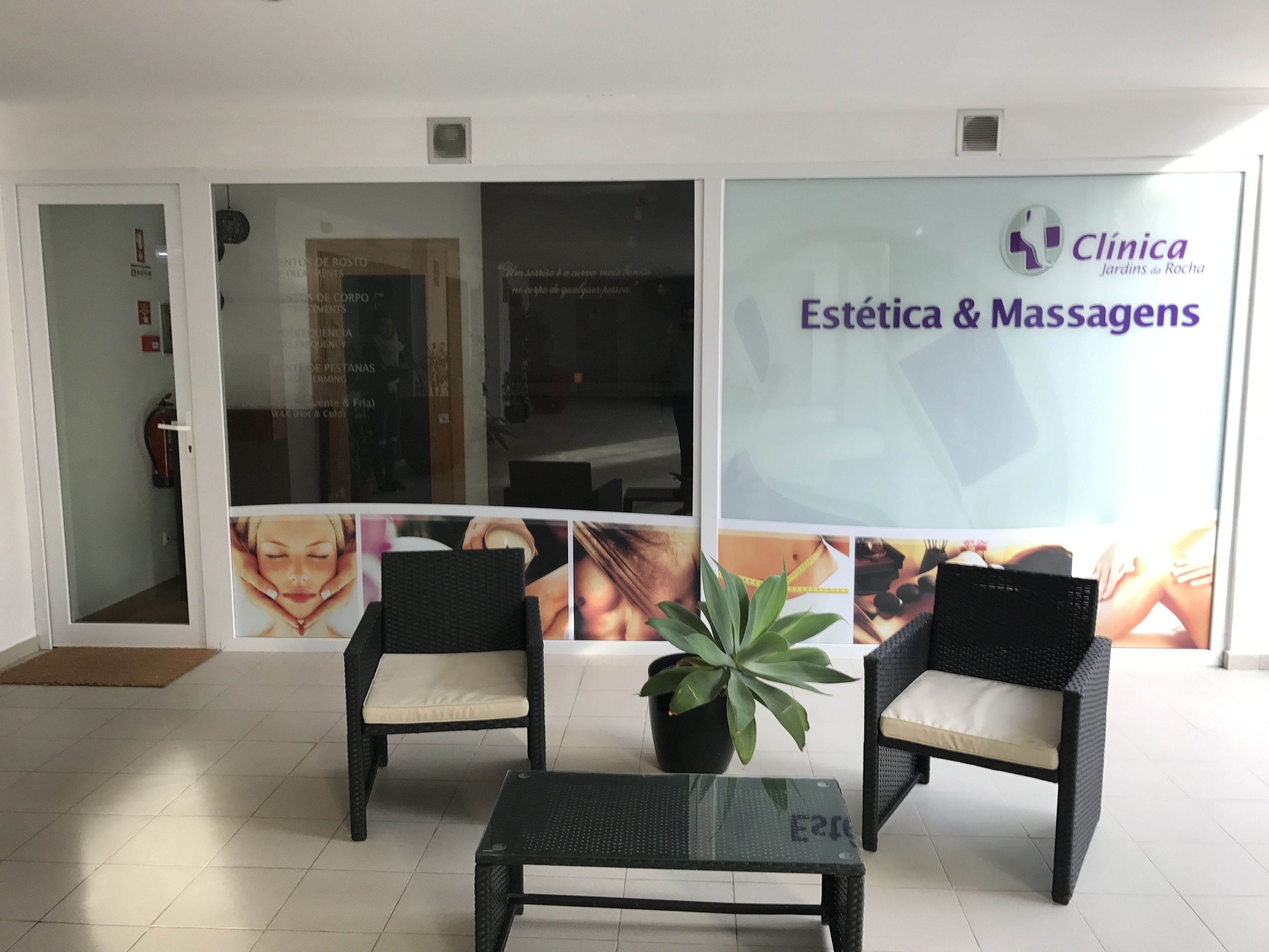 A reception area with two wicker chairs, a glass coffee table, and a potted plant. A large poster on the wall advertises 'Estética & Massagens' services.