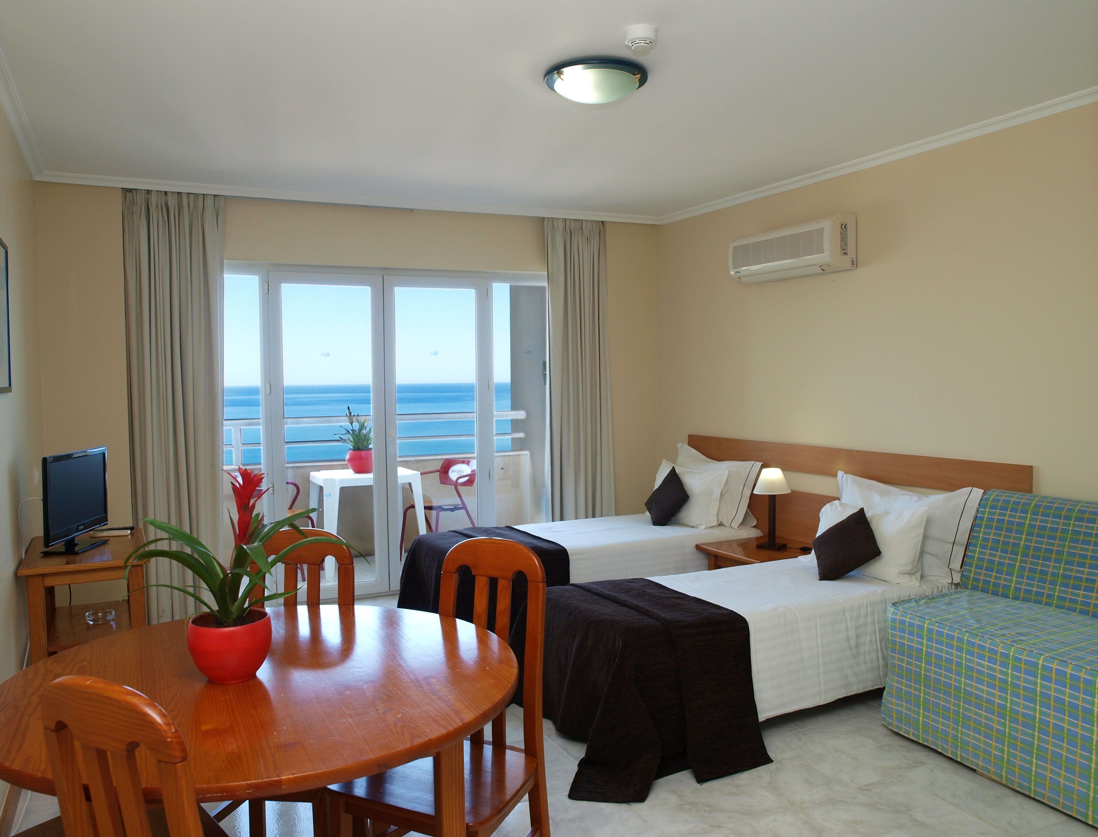 A hotel room with a bed, a sofa, a dining table with chairs, a TV, and a view of the sea.