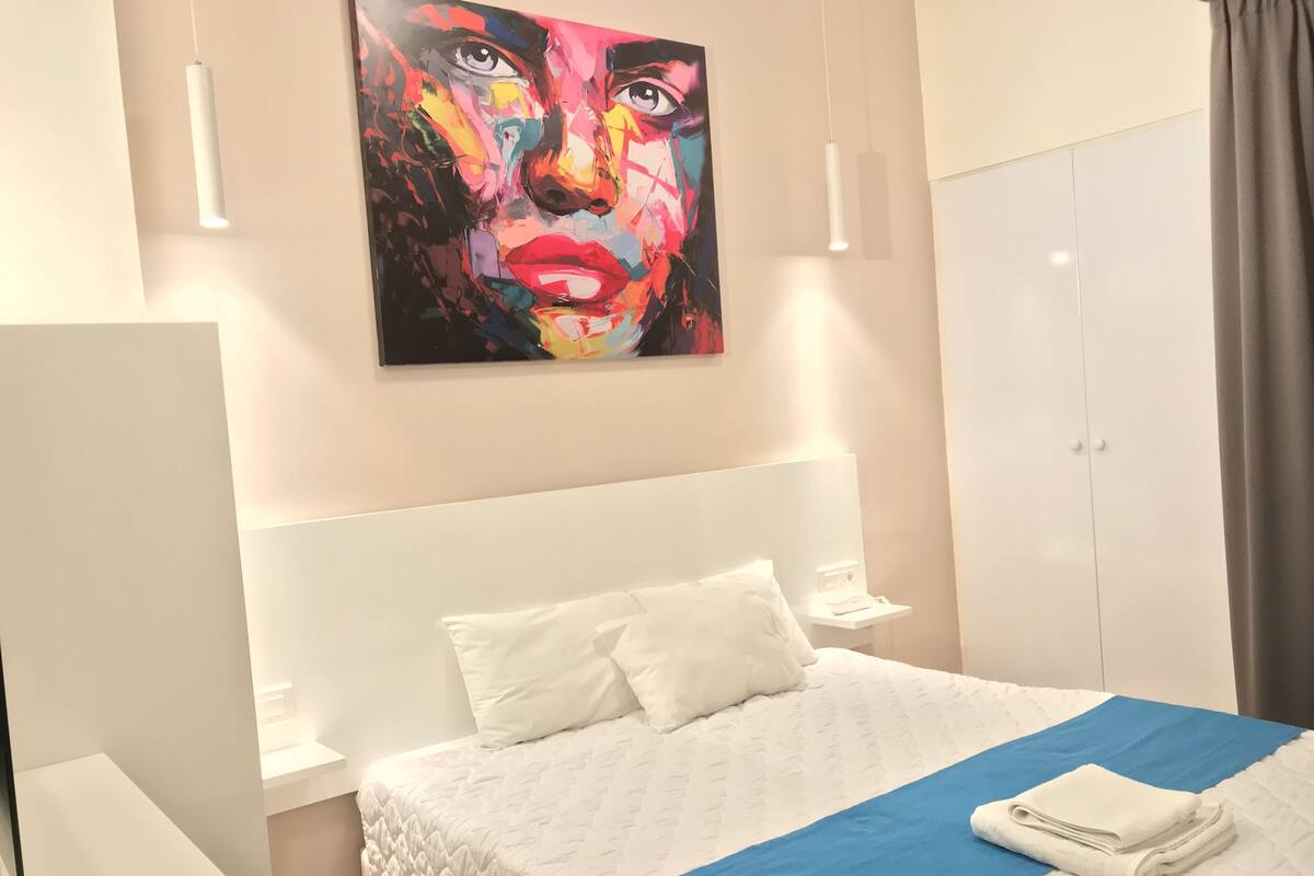 Ein modernes Hotelzimmer mit einem großen Bett, einer weißen Kopfteil und einem farbenfrohen, abstrakten Gemälde darüber.