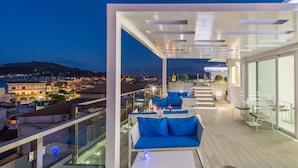 A rooftop terrace with a view of the city at dusk, featuring outdoor seating and a glass railing. - Diana Hotel (Zakynthos)