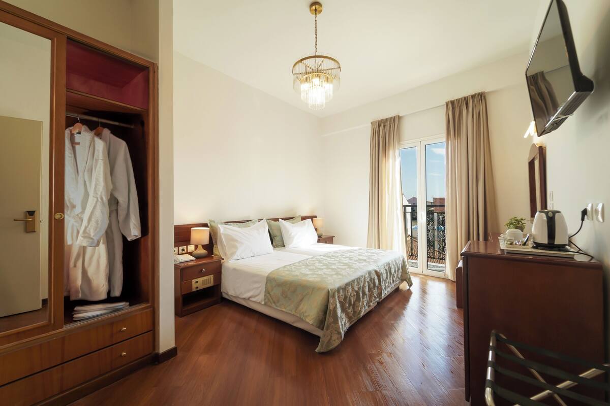 Ein Hotelzimmer mit Bett, Kleiderschrank, Schreibtisch, Fernseher und einem Balkon mit Vorhängen.