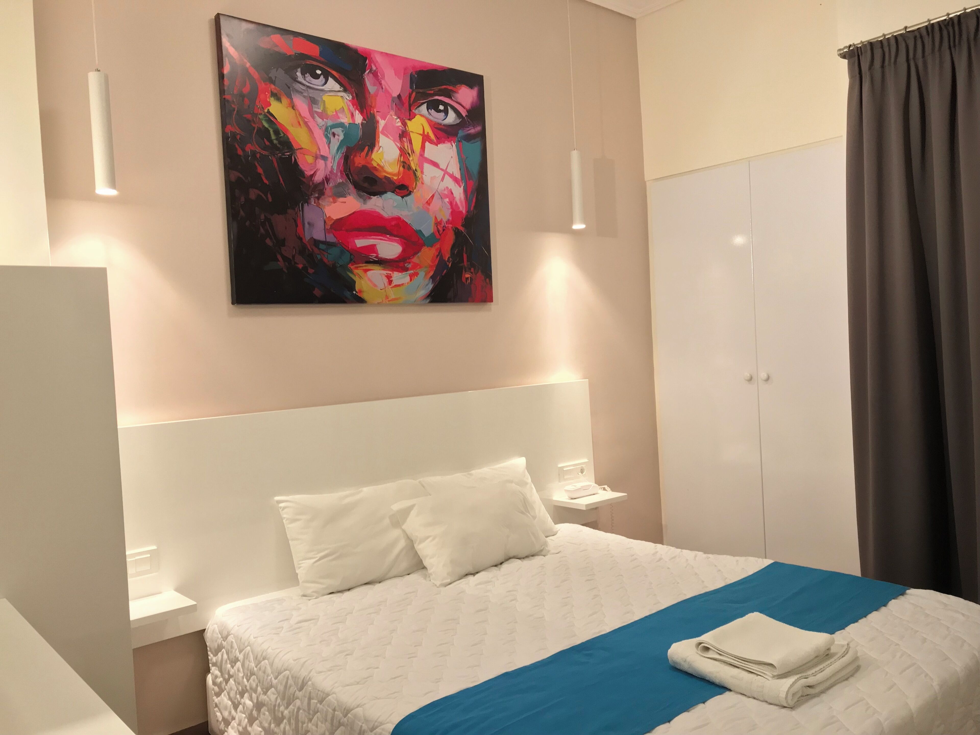 A hotel room with a large bed, white headboard, and a colorful painting above it. There is a wardrobe and a curtain on the right side.