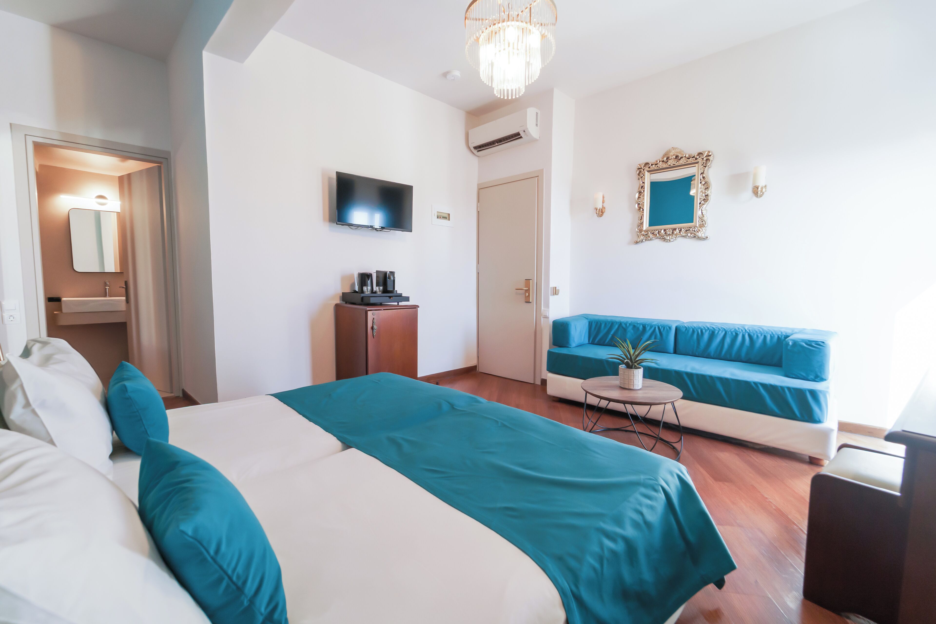 A modern hotel room with a bed, a sofa, a television, a small table with a plant, and a chandelier.
