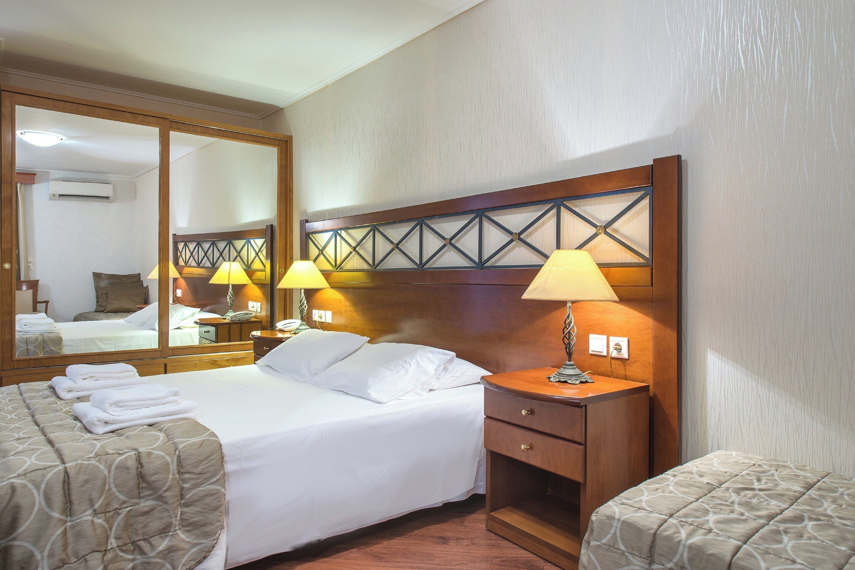 Habitación de hotel con una cama grande, dos mesitas de noche con lámparas, un espejo y un cabecero de madera.