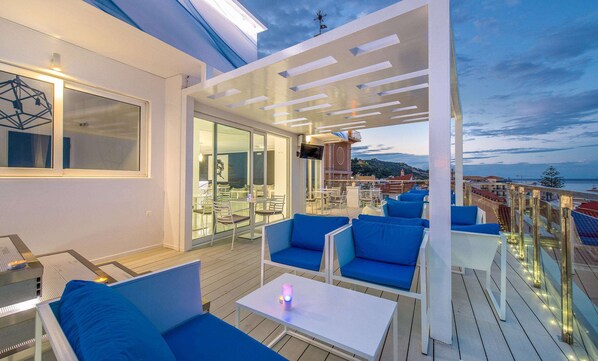 A rooftop terrace with blue seating, a TV, and a view of the city and sea. - Diana Hotel (Zakynthos)