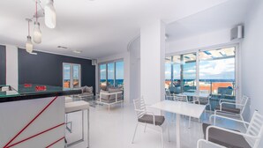A modern, well-lit room with a large window offering a view of the city. There is a white table with chairs, a bar area, and a seating area with couches. - Diana Hotel (Zakynthos)