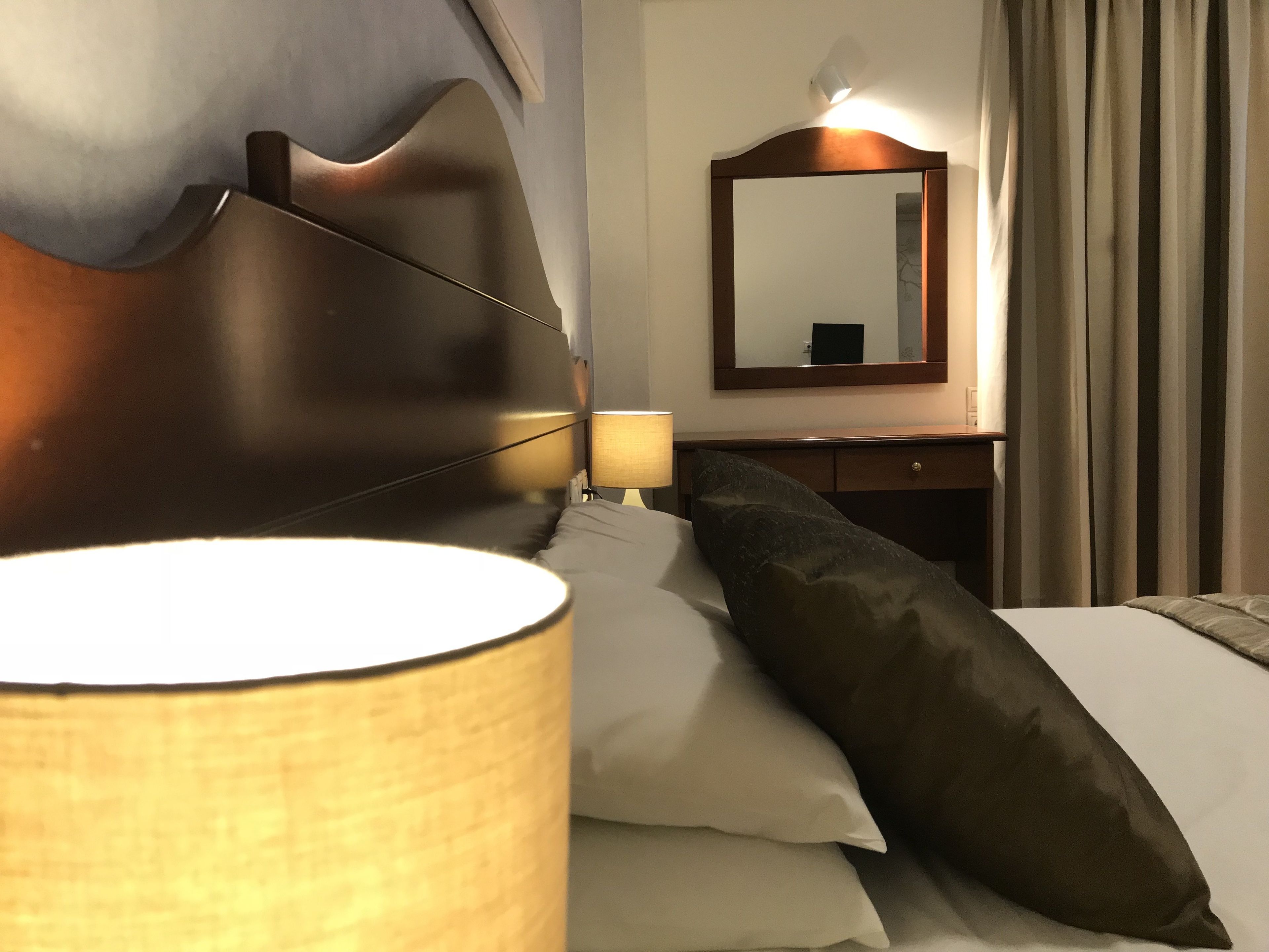 Une chambre d’hôtel avec une tête de lit en bois, une lampe de chevet, un miroir, un bureau et un lit avec des oreillers.