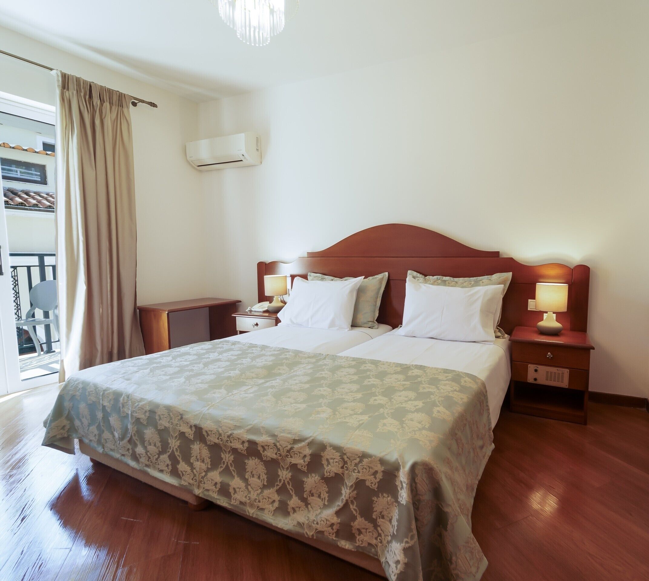 Un dormitorio de hotel con una cama grande, dos mesitas de noche con lámparas, un ventilador de techo y vistas al exterior a través de una ventana con cortinas.