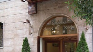 A hotel entrance with a stone facade, an arched doorway, and two potted evergreen trees. - Hotel Eden (Naples)