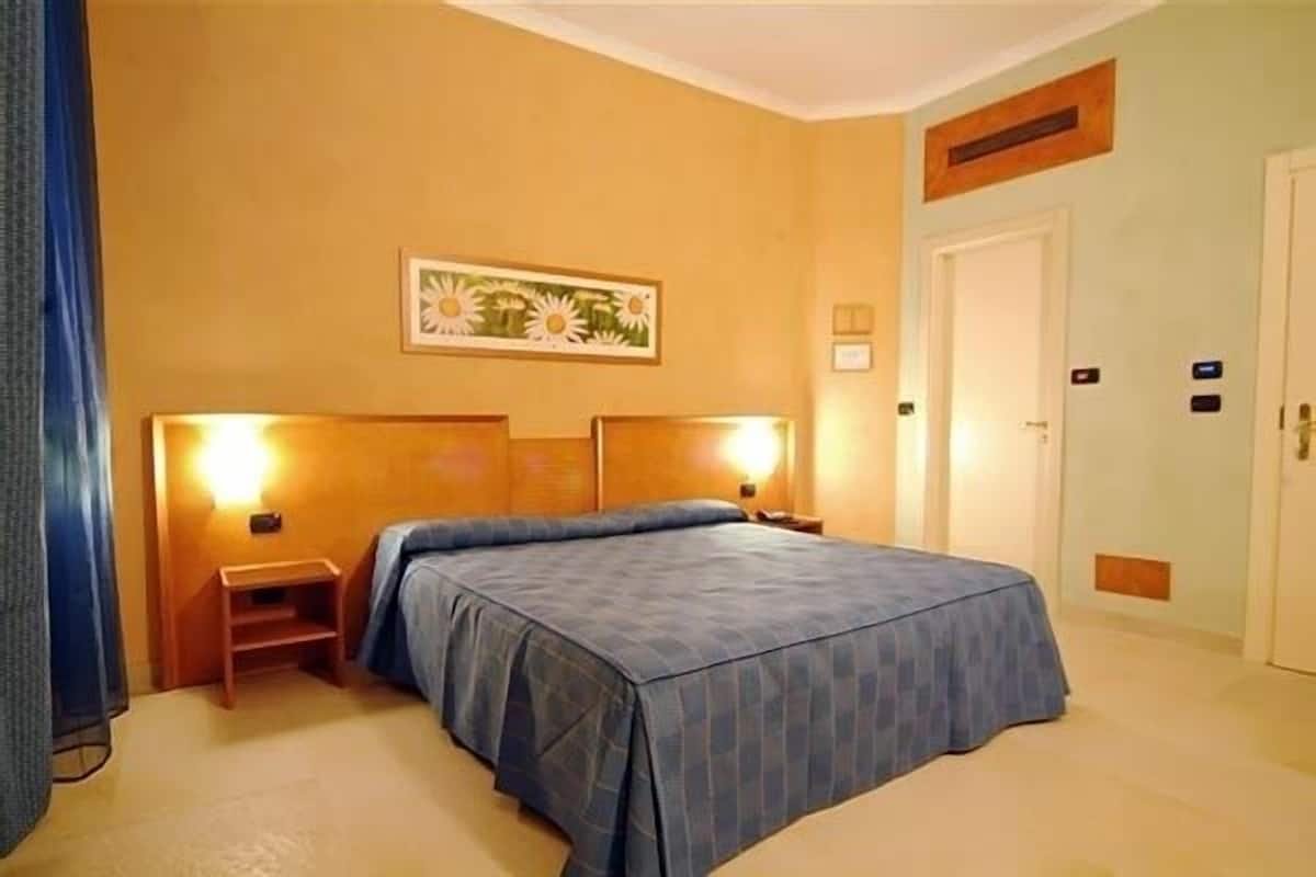 A hotel room with a bed, nightstands, a painting on the wall, and a door with an intercom.