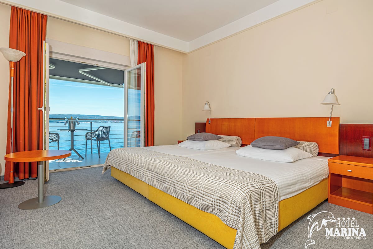 Habitación de hotel con cama, una mesita, una mesita de noche y vista al mar a través de una puerta corrediza de vidrio.