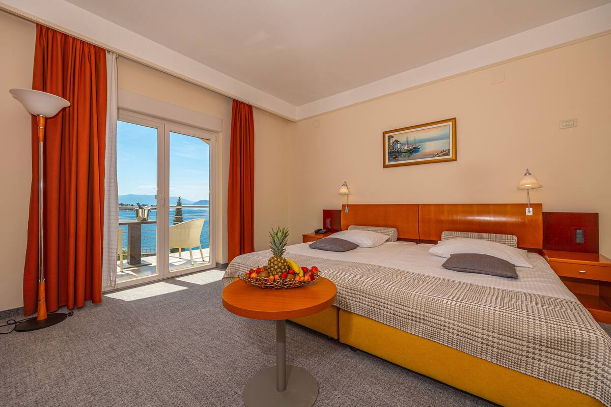 Habitación de hotel con cama, una mesa redonda con fruta, vista al mar y un cuadro en la pared.