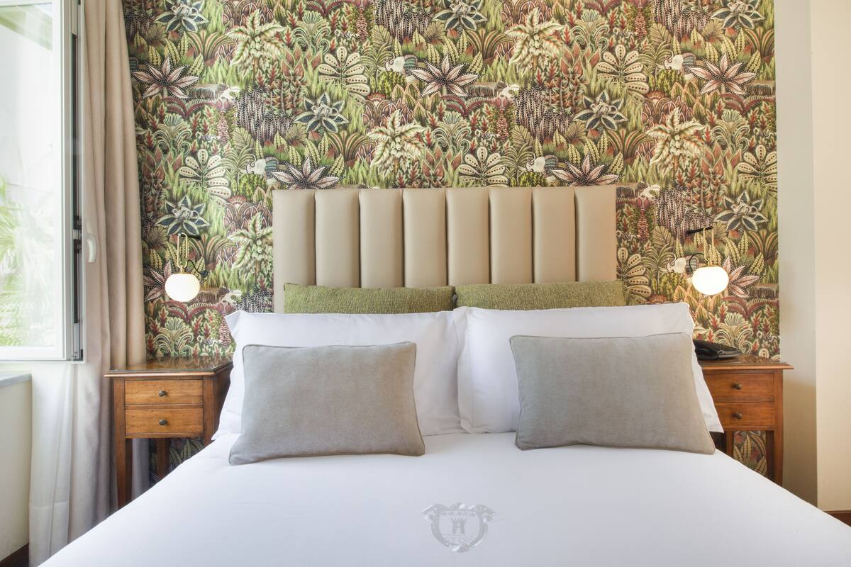 Una cama bien hecha con papel tapiz floral, dos mesitas de noche con lámparas y una ventana con cortinas.