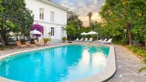A swimming pool with lounge chairs and umbrellas, surrounded by lush greenery and a white building. - Relais Villa Savarese (Sant'Agnello)