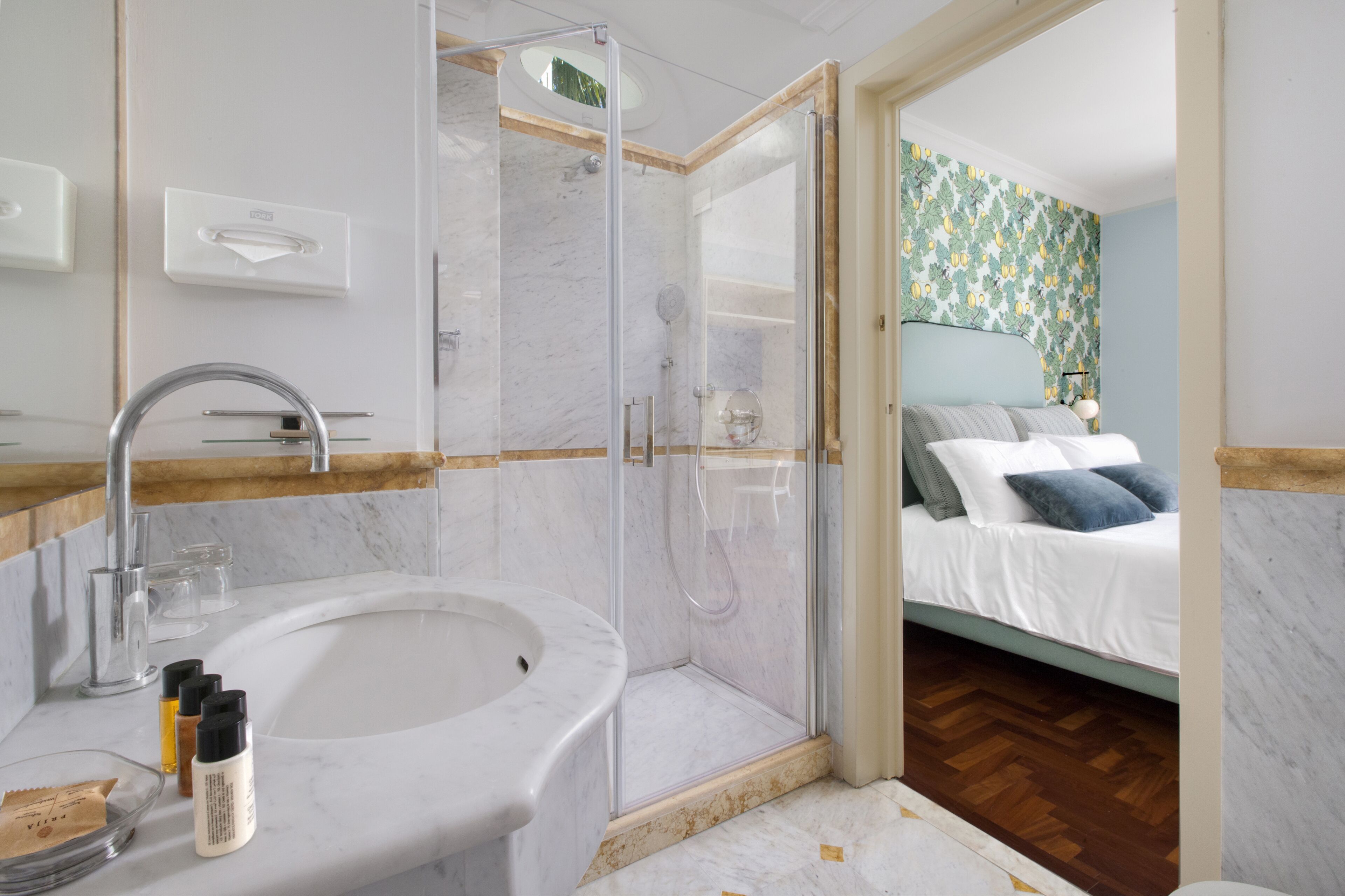 A modern bathroom with a marble sink, glass shower enclosure, and a bed with a floral wallpaper in the adjacent room.